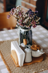wooden napkin holder with white napkins and a glass vase with dry flowers, which is on a beige checkered tablecloth on a table, next to which is a wooden chair