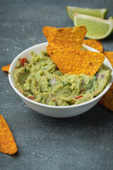 White bowl of traditional Mexican guacamole with nachos on grey concrete background. Tortilla chips with guacamole sauce dip and ingredients: avocado, cilantro, onion, hot pepper and lime