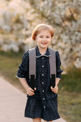 A smiling blonde girl 7-8 years old in a school uniform walks through the park with a backpack to...