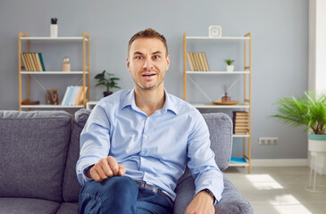 Portrait of a young man in a blue shirt and jeans sitting on the sofa at home and looking at the...