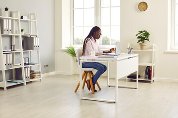 Side view portrait of a confident young business african american woman wearing casual clothes sitting at the desk on her workplace and working on a laptop at the modern white office.