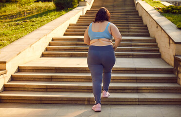 Back view of a overweight fat woman running up the stairs in the summer city park. Plus size girl wearing sportswear jogging outdoors. Weight loss, body positive, sport and fitness lifestyle concept. - obrazy, fototapety, plakaty