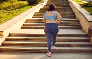 Deurstickers Back view of a overweight fat woman running up the stairs in the summer city park. Plus size girl wearing sportswear jogging outdoors. Weight loss, body positive, sport and fitness lifestyle concept. © Studio Romantic