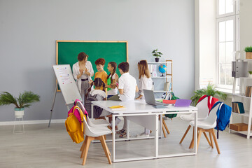 Happy children at private school take turns answering tasks of young teacher near the blackboard. Cool school room in modern style with large communal table and chairs with hanging bright backpacks.