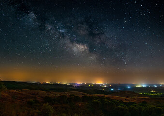 Nocturno com Via l&aacute;ctea com luzes de vila no horizonte
