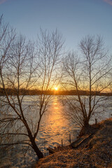 a frozen lake, on the shore of which there are trees and a wooden structure for fishing, against the backdrop of a blue sky with a bright setting sun, during the golden hour