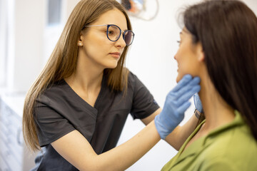 Young doctor dermatologist or cosmetologist examining facial skin of female patient in medical office