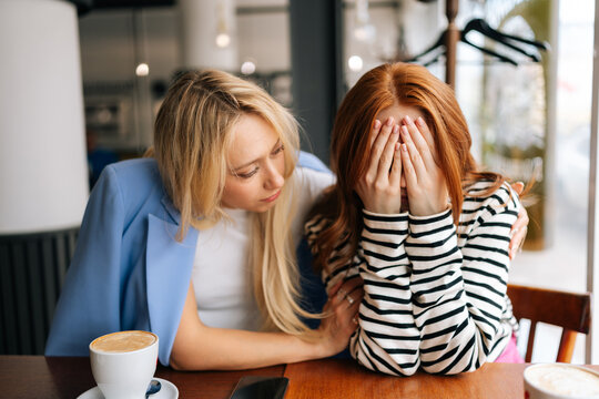 Portrait of displeased young woman and best female friend trying to comfort and cheer up sitting together in cafe by window. Unhappy redhead lady covering face with hands and crying.