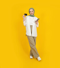 Online shopping, full size young caucasian muslim woman in hijab holding credit card and mobile phone online shopping concept idea image. Isolated yellow background. People islam religious lifestyle.