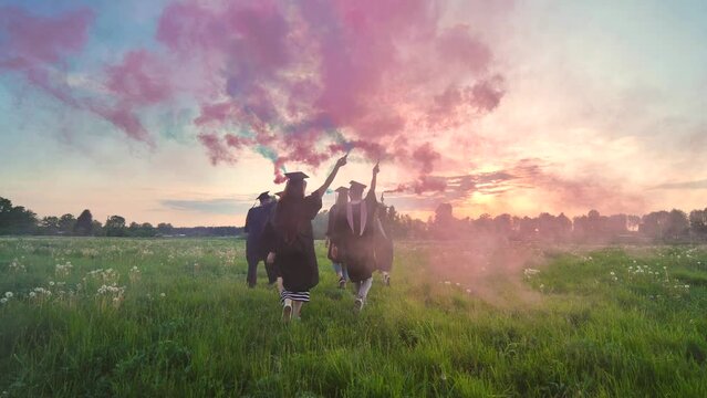 Graduates in costume walk across a field with coloured red and blue smoke at sunset.