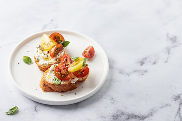Bruschetta sandwiches with tomatoes, cream cheese, olive oil and basil on a plate on white marble background, copy space. Traditional italian antipasti