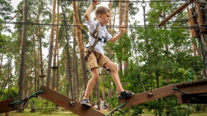 Young boy holding ropes and trying to cross the wobbly bridge in adventure park at forest