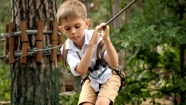 Portrait of little boy holding tight safety cable while walking on rope bridge at extreme adventure park. Kids sports, summer holiday, fun outdoors, scouts.