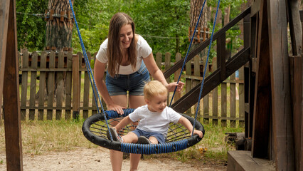 Young smiling mother swinging her cute toddler boy in rope nest swing at park playground