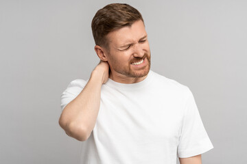 Man suffers from neck pain. Painful ache in neck of exhausted middle aged man touching on occiput on grey studio background. Symptom of cervical chondrosis, hernia of spine, inflammation of vertebra.