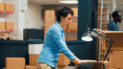 Female entrepreneur putting products in cardboard boxes, shipping merchandise to clients. Young person preparing shipment with storehouse goods, retail work and logistics. Tripod shot.