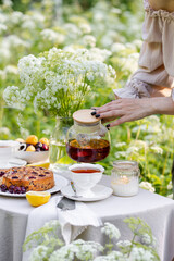 Summer elegant picnic. Woman in white dress purring tea from teapot at wooden table in garden. Beautiful dishwater, white porcelain, tablecloth, homemade cherry pie.