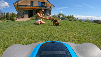 POV = POINT OF VIEW: Autonomous lawnmower approaching playful woman and her dog - obrazy, fototapety, plakaty