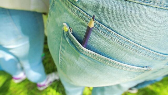 Close-up of purple pencil sticks out of the back pocket of a woman's blue jeans, standing on the grass on a summer day