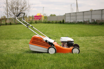 side view of modern orange-grey electric lawn mower cutting bright lush green grass. Gardening work tools. Rotary lawn mower machine on lawn. Professional lawn care service. Place for text