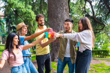 Multiethnic group of birthday party in the city park hugging and toasting with the plastic cups