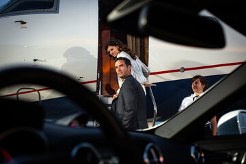 Successful couple boarding private Jet , man looking back over shoulder on tarmac of airport, view from drivers seat of sports car parked next to plane on tarmac 