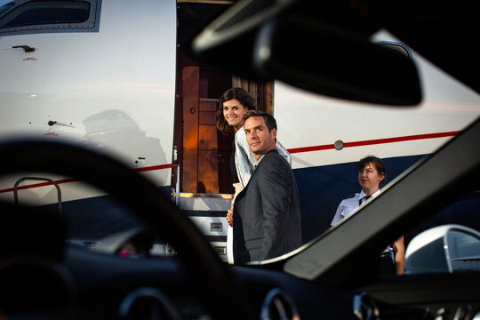 Successful couple boarding private Jet looking back over shoulders on tarmac of airport, view from drivers seat of sports car parked next to plane on tarmac 