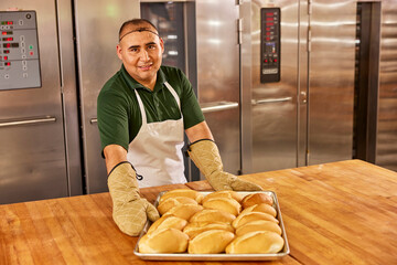 Portrait of hispanic smiling baker in a commercial kitchen of grocery placing tray of freshly baked bolillos from oven on to large worktable.