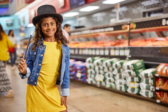 Young girl with magicians top hat and wand standing in isle of grocery store, meat market in the background 