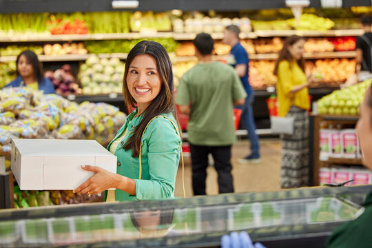 Female customer with custom cake in box leaving bakery counter in grocery store, customers in produce section in background 