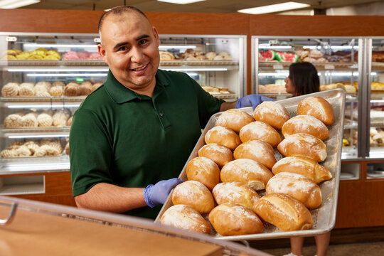 Portrait of Grocery store baker, smiling to camera refilling bin with tray of fresh baked Bolillos, a type of savory bread made in Mexico and Central America.