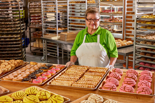 Portrait of female baker with apron and hair net in commercial kitchen of grocery store with large selection of freshly baked sweet breads surrounding her 