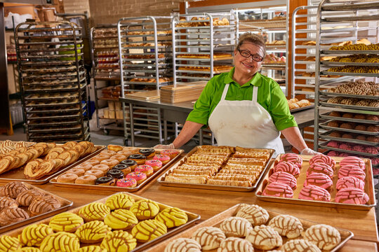 Portrait of smiling female baker with apron and hair net in commercial kitchen of grocery store standing with large selection of freshly baked sweet breads