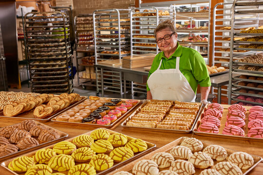 smiling female baker with glasses, apron and hair net in commercial kitchen standing behind trays of freshly baked sweet breads
