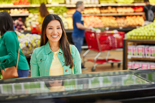 Female customer smiling looking into display case of bakery counter in grocery store, customers in produce section in background 