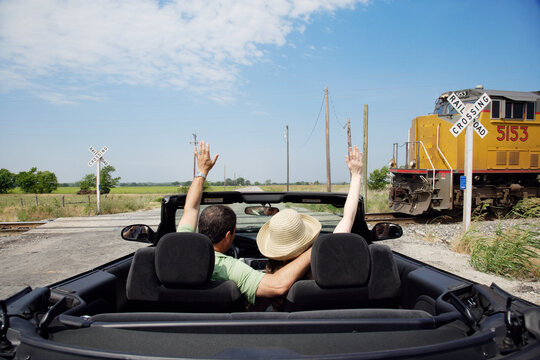 Middle aged baby boomer couple driving down country road in convertible with top down, Stopped at a railroad crossing waving to passing train 
