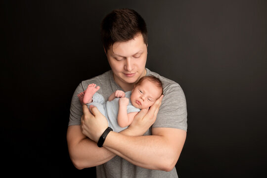 A cute newborn baby boy sleeps in a blue overalls in the first days of life. In the arms of the father. Warm parental hugs. On a black background. Professional portrait photography.