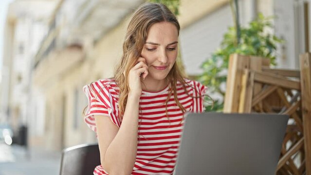 Young blonde woman talking on smartphone using laptop sitting on table at coffee shop terrace