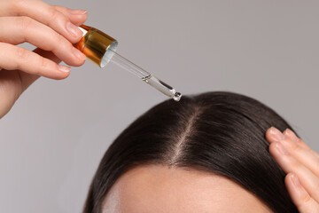 Woman applying essential oil onto hair roots on light grey background, closeup