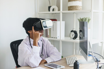 Young Asian doctor siiting on workdesk wearing virtual glasses in virtual reality in clinic. Man in...