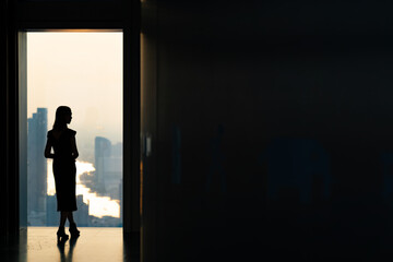 Obraz na płótnie Canvas Silhouette Asian woman standing by the window at skyscraper building in metropolis and looking cityscape at summer sunset. Beautiful girl enjoy urban lifestyle activity in the city on holiday vacation