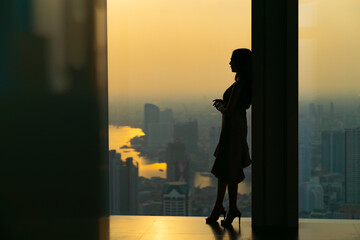 Silhouette Asian woman standing by the window at skyscraper building in metropolis and looking...
