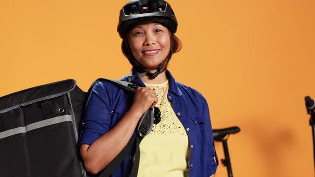 Portrait of happy asian courier using bike and thermic bag to transport perishables to customer. Delivery part time employee isolated over orange studio background wearing safety gear, close up