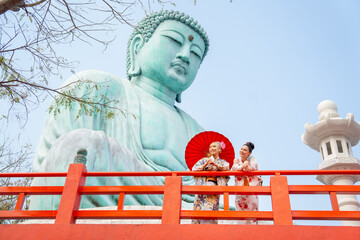Fototapeta premium Wide shot of two Asian pretty women stay close to red railing in the area of base of green big buddha statue and they look to left side with happiness.