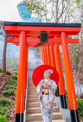 Beautiful woman with Japanese style dress hold red umbrella and stand on base stairs with red arched entrance in area of tourist attraction in Thailand.