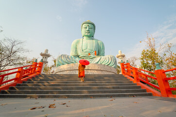 Wide shot back of young Asian monk hold red umbrella and walk up on stair in front of green big...