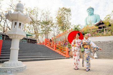 Fototapeta na wymiar Wide shot of two Asian women wear japanese style dress with one hold red umbrella and stand in front of stair to green big buddha statue.