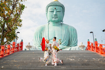 Two Asian young woman with japanese style dress stay on stair in front of green big buddha statue...