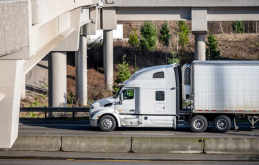 Side view of the white bonnet big rig semi truck with refrigerator semi trailer running on the highway road under the bridge and overpass intersection