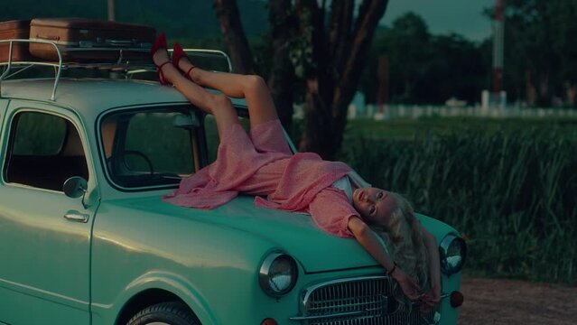 A blonde in a pink vintage dress lies on the hood of a turquoise-colored retro car